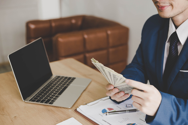 Man in a business suit sitting at a table with a laptop and a fan of paper money.